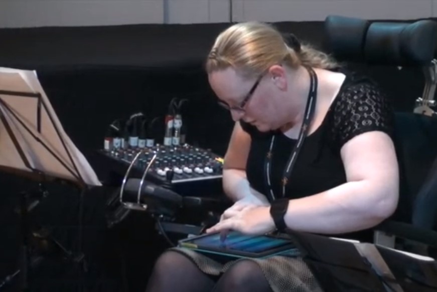 A blonde lady in a powered wheelchair is playing music on an iPad with a mixer and a music stand